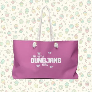May include: A large, pink tote bag with white rope handles. The bag features the text "I AM JUST A DUNGJANG GIRL" in white block letters, with small bow graphics. The background is a pattern of cupcakes and ice cream cones.