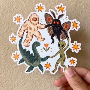 May include: A die-cut sticker featuring a beige Bigfoot, a black moth-like creature with orange eyes, a green aquatic monster, and a grey alien holding hands in a circle. The design includes orange star shapes and a small orange flower in the center.