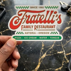 May include: A white sticker with the text "Fratelli's FAMILY RESTAURANT" in red and brown. The sticker also includes the text "ASTORIA • OREGON" and "PIZZA ~ ICE CREAM ~ WATER ~ TONGUE". The sticker has a green and white checkered border and the text "SINCE 1985".