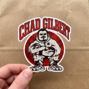 May include: A die-cut sticker featuring a cartoon illustration of a person with a goatee wearing a hooded sweatshirt and sneakers, arms crossed, set against a red circular background with the text CHAD GILBERT arched in red capital letters.