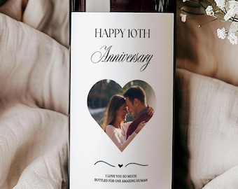 May include: A wine bottle with a custom label for a 10th anniversary. The label features a heart-shaped photo of a couple and the words "Happy 10th Anniversary". The bottle is dark with a brown top, set against a neutral fabric and white flowers.