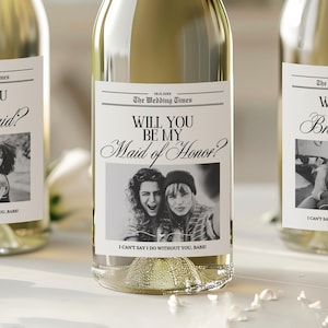 May include: Three champagne bottles with labels saying "Will You Be My Bridesmaid?" and "Will You Be My Maid of Honor?" The labels have black and white photos. The bottles have gold foil tops and are filled with a light-coloured liquid.