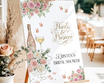 May include: A white rectangular sign on a wooden easel featuring watercolor illustrations of pink roses, butterflies, a bottle of prosecco, a glass, and pearl strands. The text reads Pearls & Prosecco and Kristen's BRIDAL SHOWER.