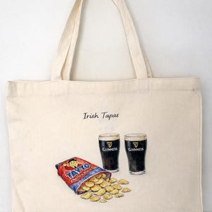 Könnte beinhalten: Cremefarbene Tragetasche mit dem schwarzen Aufdruck "Irish Tapas". Die Tasche zeigt eine Illustration von zwei Guinness-Gläsern und einer Tüte Tayto-Chips. Die Tasche hängt an einem kupferfarbenen Haken.