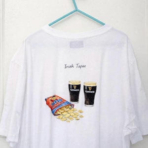 Könnte beinhalten: Weißes T-Shirt mit einer Illustration von zwei Pints Guinness, einer Tüte Tayto-Chips und dem Text "Irish Tapas". Das T-Shirt hängt an einem blauen Kleiderbügel.