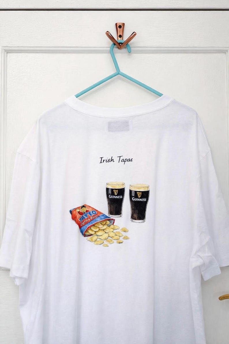 May include: White t-shirt with an illustration of two pints of Guinness, a bag of Tayto crisps, and the text "Irish Tapas." The shirt is hanging on a blue hanger.