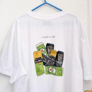 Pu&ograve; includere: T-shirt bianca con un'illustrazione ad acquerello di varie lattine di birra e sidro, tra cui Bulmers e Guinness. Il testo "BIG BAG OF CANS" &egrave; stampato sopra la grafica. La maglietta &egrave; appesa a una gruccia blu.