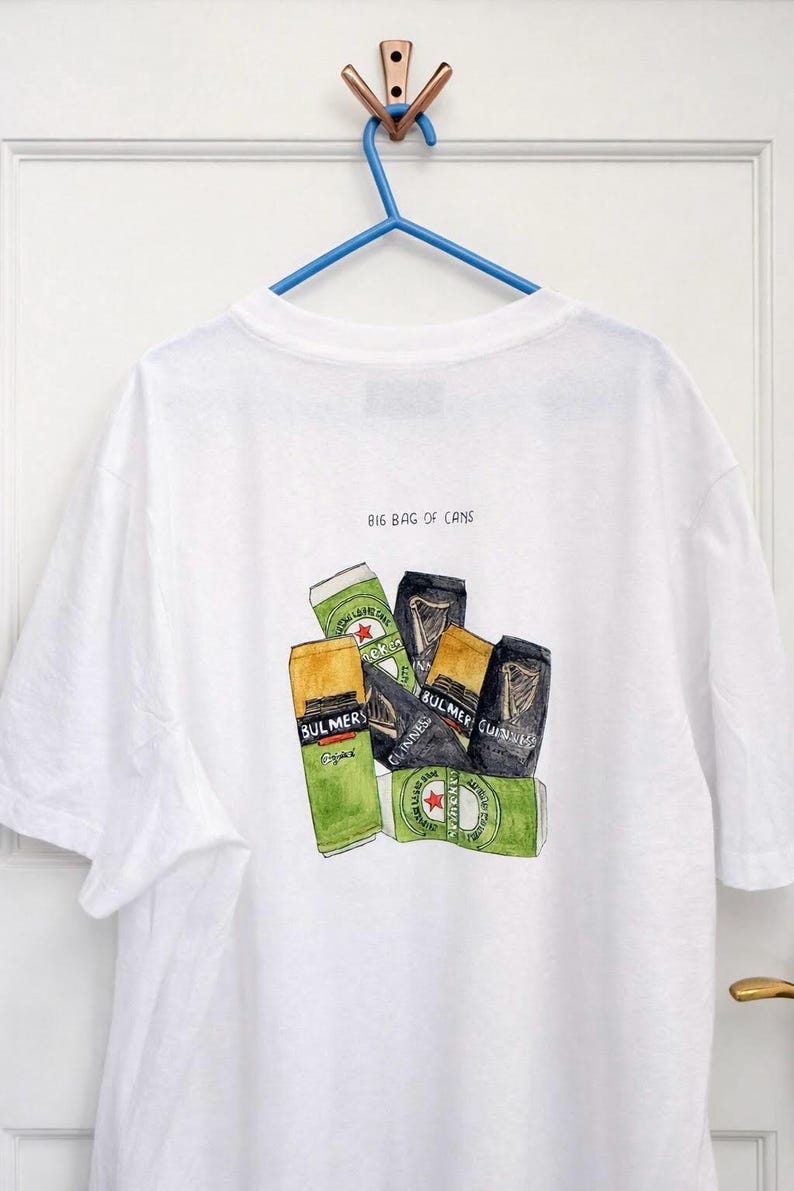 Pu&ograve; includere: T-shirt bianca con un'illustrazione ad acquerello di varie lattine di birra e sidro, tra cui Bulmers e Guinness. Il testo "BIG BAG OF CANS" &egrave; stampato sopra la grafica. La maglietta &egrave; appesa a una gruccia blu.