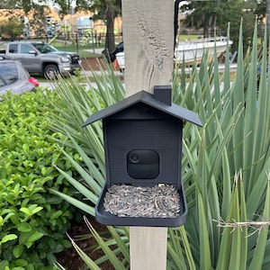May include: A black bird feeder with a house-like design, mounted on a wooden post. The feeder is filled with sunflower seeds and has a camera. The background features green plants and a glimpse of a vehicle.