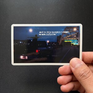 May include: A rectangular sticker featuring a night scene of a person standing on a dark truck with arms outstretched. The text "and in this moment I swear, we are infinite." is printed in white. A bridge structure is visible in the background.