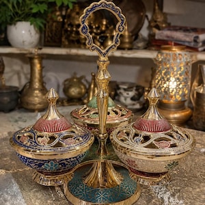 Op de afbeelding: Een goudkleurige serveerstandaard met drie niveaus en drie decoratieve kommen. De kommen hebben rode, blauwe en groene accenten, met ingewikkelde bloempatronen en opengewerkte ontwerpen. De standaard heeft een handvat met een blauw en goud filigraan ontwerp.