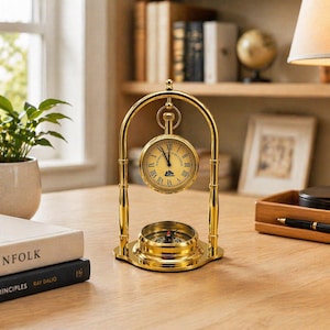 Puede incluir: Un juego de reloj y brújula de escritorio de color dorado. El reloj tiene una esfera redonda con números romanos y una pequeña brújula en la base. El conjunto es de metal y está sobre un escritorio de madera.