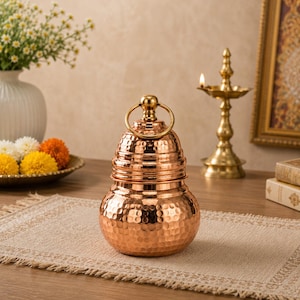 May include: A copper-colored, hammered metal container with a rounded base and a bell-shaped top. It features a gold-colored ring handle and a decorative, textured surface. The container sits on a beige placemat, suggesting a decorative or ceremonial purpose.