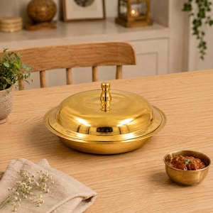 May include: A gold-colored serving dish with a lid and a small bowl of food on a wooden table. The serving dish has a round shape with a lid and a small handle on top. The small bowl is also gold-colored and contains a red sauce.