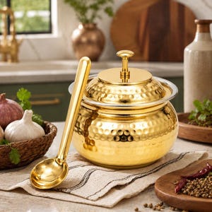 May include: A polished gold-tone metal serving pot with a matching lid and a long-handled gold-tone serving spoon. The pot features a rounded body with a hammered texture and a small knob handle on the lid, providing a reflective metallic finish.
