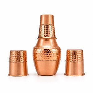 Peut inclure: Un service à boissons de couleur cuivre, comprenant une carafe haute à texture martelée avec un couvercle et deux tasses assorties. La carafe a une base bombée et un col plus étroit. Les tasses sont cylindriques avec une texture martelée.