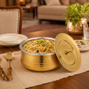 Puede incluir: Un cuenco de servicio redondo de metal martillado dorado lleno de biryani, cubierto con anacardos y hierbas. El cuenco cuenta con una tapa extraíble a juego con un pomo central y se acompaña de una cuchara de servir de mango largo.