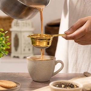 Puede incluir: Un colador de té dorado se utiliza para verter té en una taza de cerámica moteada. El té es de color marrón cálido y el colador tiene una malla fina. La taza está sobre una superficie de madera.
