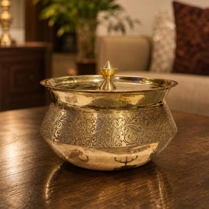 May include: A gold-colored metal bowl with a lid, featuring an ornate, engraved floral pattern around the midsection. The bowl has a rounded shape and sits on a wooden surface. The lid is topped with a small, pointed finial.