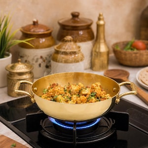 May include: A golden brass pan with two handles, filled with cooked cauliflower and herbs, sits on a lit gas stove. Steam rises from the food. Background includes ceramic jars, a small plant, and a basket.