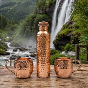 May include: A copper water bottle and two copper mugs with a hammered texture are displayed on a wooden surface. The bottle is tall and cylindrical with a screw-on cap. The mugs have handles. A waterfall and lush greenery are in the background.