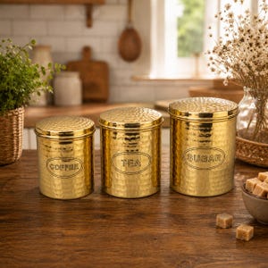 Könnte beinhalten: Ein Set aus drei zylindrischen, goldfarbenen Metalldosen mit gehämmerten Oberflächen und passenden Deckeln. Jede Dose hat ein ovales Etikett mit dem schwarzen Schriftzug COFFEE, TEA oder SUGAR, angeordnet nach ihrer Größe.