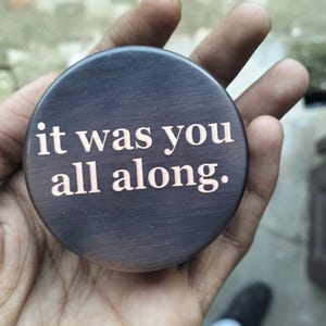 May include: A round, dark brown object with the phrase "it was you all along." in a lighter color. The object has a brushed metal appearance and is held in a hand.