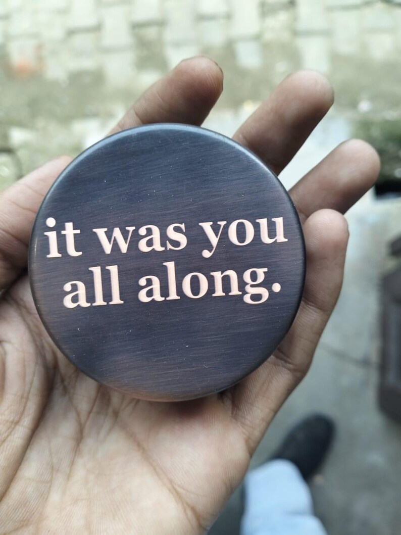 May include: A round, dark brown object with the phrase "it was you all along." in a lighter color. The object has a brushed metal appearance and is held in a hand.