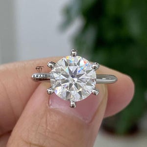 May include: A silver ring featuring a large, round, clear gemstone. The gemstone is faceted, reflecting light and creating a sparkling effect. The ring has a simple band and six prongs securing the stone. The logo "KRAFT JEWEL" is visible.