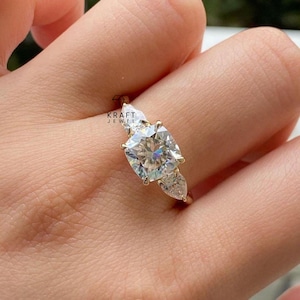 Peut inclure: Une bague en or ornée d'une grosse pierre centrale de taille carrée, flanquée de deux pierres en forme de poire. La bague est sertie de multiples facettes, créant un effet scintillant. La bague est tenue par une main.