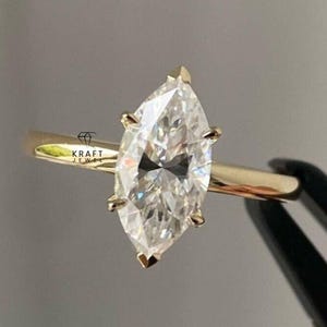 May include: A marquise-cut diamond engagement ring with a gold band. The diamond is the focal point, set with six prongs. The ring is held by a black tool against a grey background. The logo "KRAFT JEWEL" is visible.