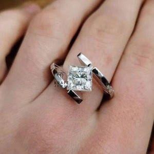 May include: A silver ring featuring a square, clear gemstone. The ring has a distinctive design with geometric elements. The ring is on a person's finger. The text "KRAFT JEWEL" is visible.
