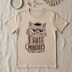 Könnte beinhalten: Ein beiges Kurzarm-T-Shirt mit einer grafischen Illustration einer mürrischen braunen Katze mit einer Haarsträhne auf dem Kopf. Unter der Katze steht der Text I HATE MONDAYS in schwarzen Großbuchstaben. Das Shirt hat einen Rundhalsausschnitt.