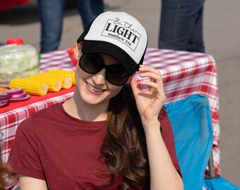 May include: A black and white trucker hat with the text "Be the LIGHT" and "matthew 5:14" is worn by a person. The person is wearing a maroon t-shirt and sunglasses. The background includes a red and white checkered tablecloth and corn on the cob.
