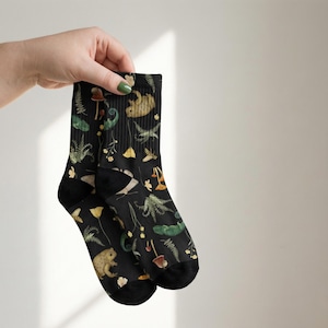 May include: Black socks with a whimsical pattern of mushrooms, ferns, frogs, and other forest elements. The socks have a dark background and a repeating design. The socks are held up by a hand.