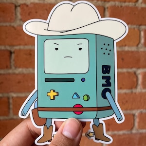 May include: A die-cut vinyl sticker featuring a light blue, rectangular, anthropomorphic video game console character wearing a white cowboy hat and brown cowboy boots. The character has a screen face, a yellow plus sign, blue triangle, green circle, red circle, and the text BMC printed vertically on the side.