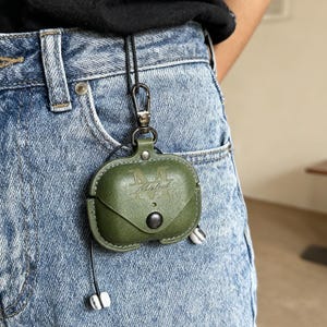 Op de afbeelding: Olijfgroene leren AirPod-hoes met een drukknoopsluiting en een metalen sluiting. De hoes is bevestigd aan een zwart koord en hangt aan een spijkerbroek. Het woord "Arbel" is in reliëf op de voorkant geplaatst.
