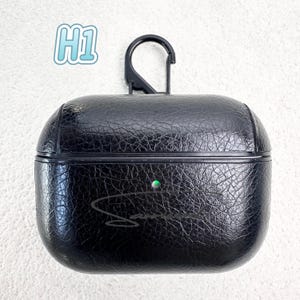 May include: Black textured leather AirPods case with a black carabiner clip. The case has a green indicator light and a signature in cursive. The text "H1" is in the upper left corner.