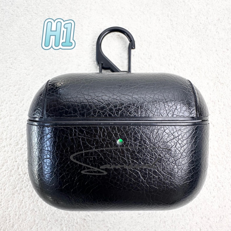 May include: Black textured leather AirPods case with a black carabiner clip. The case has a green indicator light and a signature in cursive. The text "H1" is in the upper left corner.