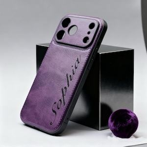 Puede incluir: Una funda de teléfono morada con el nombre "Sophia" en escritura cursiva. La funda tiene un aspecto de cuero texturizado y un borde negro. Una pequeña bola morada difusa está cerca. La funda está diseñada para un teléfono con múltiples lentes.