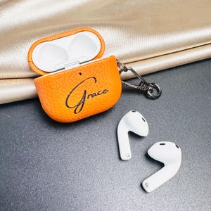 May include: Orange leather AirPod case with the word "Grace" in script. The case is open, revealing a white interior. Two white AirPods are next to the case, and a silver clasp is attached to the case.