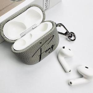 May include: Gray textured AirPods case with the name "Feng" in black script. The case is open, revealing the white interior. Two white AirPods are next to the case, along with a silver-colored carabiner clip.