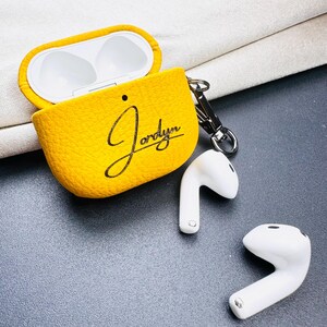 May include: Yellow textured AirPods case with the name "Jordyn" in black script. The case is open, revealing the white interior. Two white AirPods are next to the case, and a silver clasp is attached to the case.
