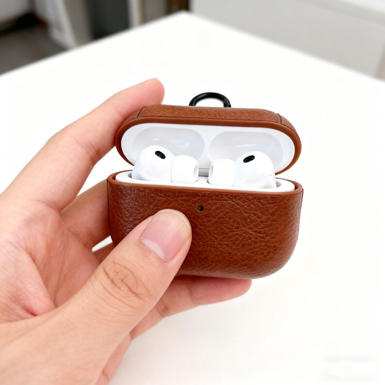 May include: Brown leather AirPod case with white AirPods inside. The case is open, revealing the earbuds. The case has a textured surface and a small black loop at the top. The background is white.