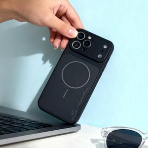 Puede incluir: Funda de teléfono negra con un círculo de carga magnética y recortes para la cámara. La funda tiene un diseño elegante con una firma en la esquina inferior derecha. La funda del teléfono se sostiene sobre un portátil.
