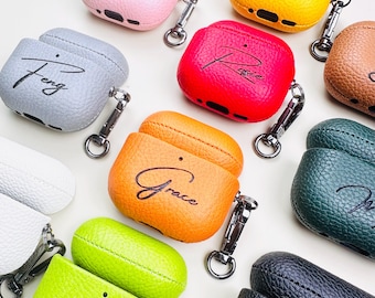May include: A selection of personalised AirPod cases in colours such as pink, yellow, red, orange, green, grey, and black. Each case has a name in cursive writing and a silver carabiner clip for attachment.