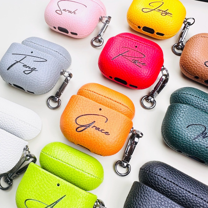 May include: A collection of personalized AirPod cases in various colors, including pink, yellow, red, orange, green, gray, and black. Each case features a name in cursive script and a silver carabiner clip for attachment.