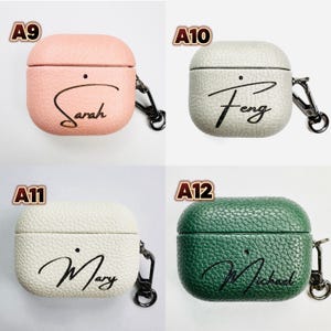 May include: Four personalized AirPod Pro cases in various colors: light pink, light gray, off-white, and dark green. Each case has a name in cursive script. The cases have a textured leather-like appearance and a silver carabiner clip.