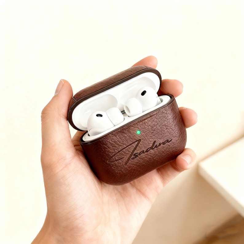 May include: A brown leather AirPods Pro case with the word "Sadiva" embossed on the front. The case is open, revealing white AirPods inside. A small green light is visible on the case. The case is held in a hand.
