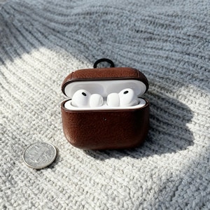 May include: Open brown leather case with white earbuds inside. A silver coin is next to the case. The case has a black loop attached to the top.